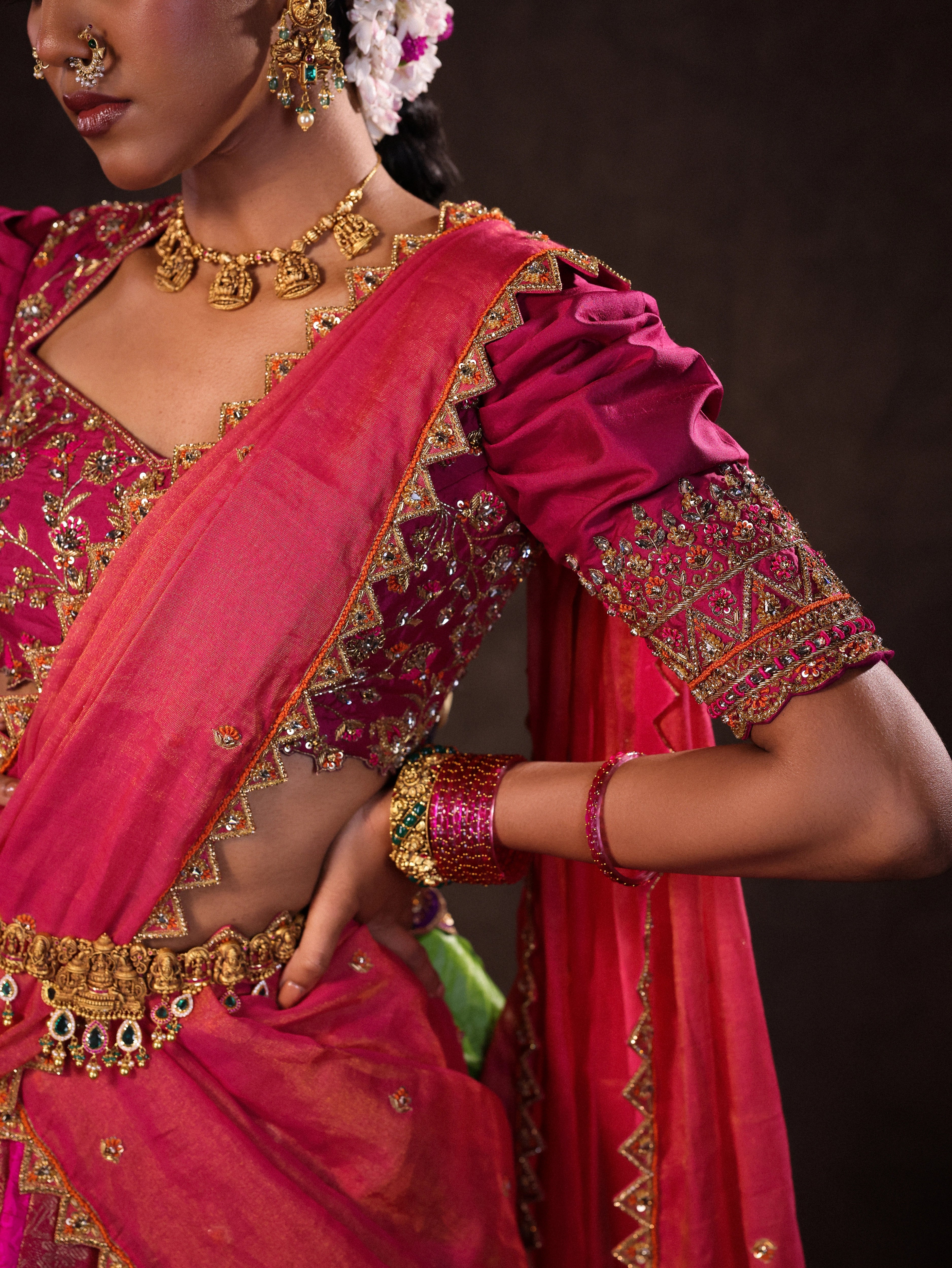 Model Gayathri Dhanasekar showcases sleeve in a pink & orange silk half saree handcrafted with zardozi, cutdana (bugle beads), resham (silk thread), sequins and rhinestones. Team Archana Karthick completed the style with pink glass bangles, a ring, a choker, jhumkas, nose ring and a maang tikka.