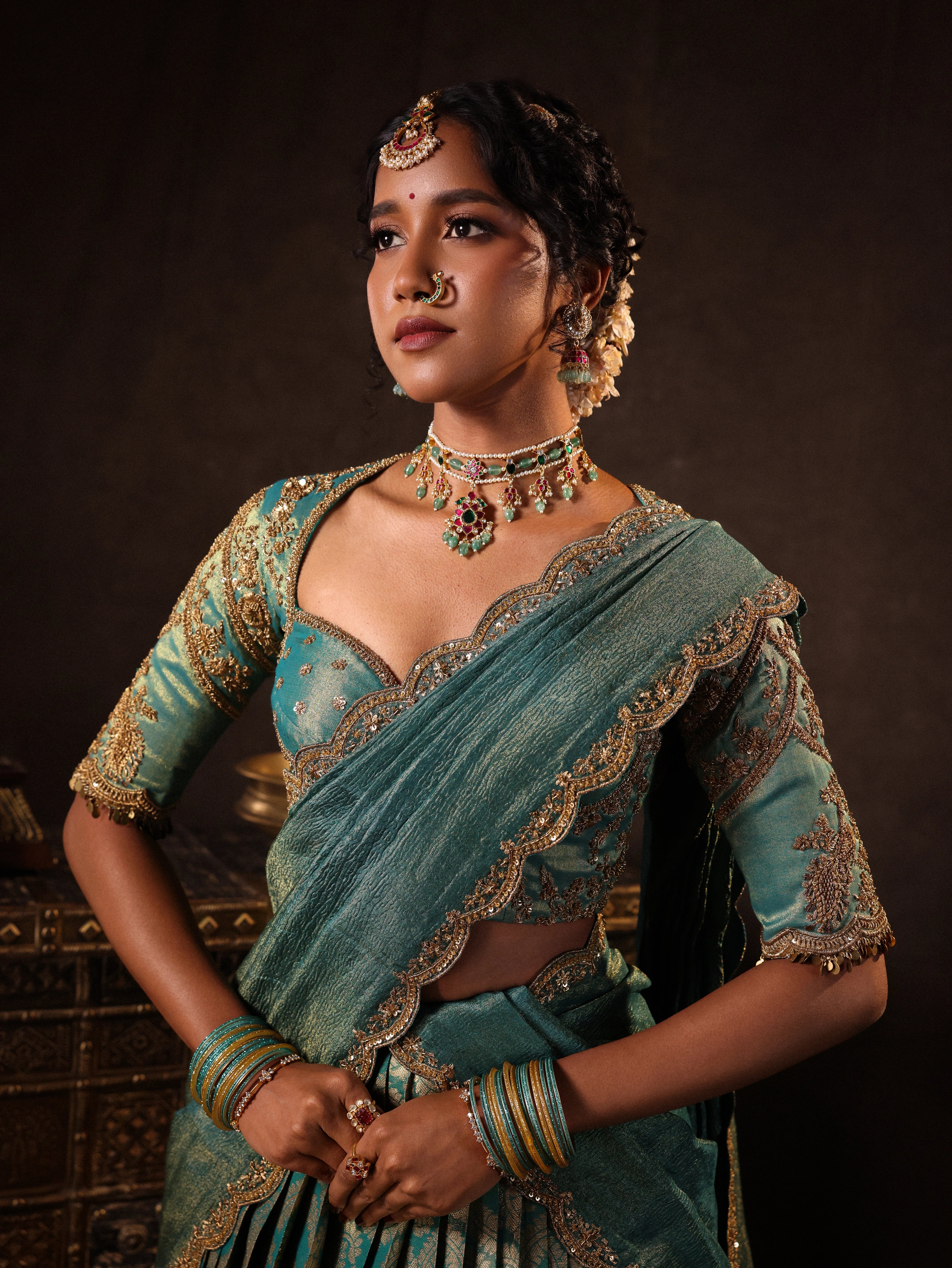 Model Gayathri Dhanasekar showcasing front profile in a teal half saree (silk blouse and skirt and tissue dupatta) handcrafted with zardozi, zari, cutdana, sequins and metal coin hangings. Team Archana Karthick completed the style with teal glass bangles, a ring, a choker, jhumkas, nose ring and a maang tikka.