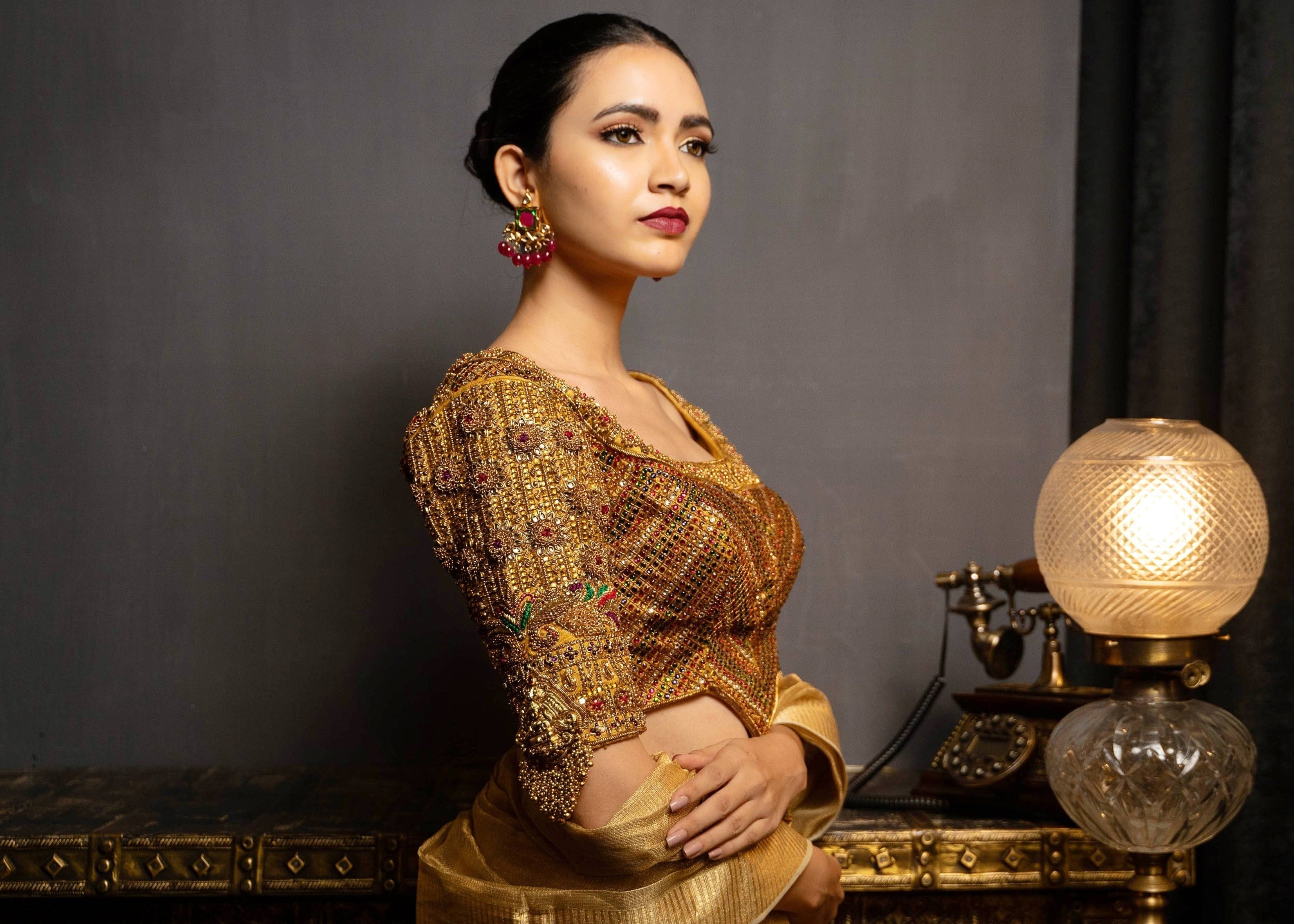 Model Preeti Kumari posing in a gold handcrafted raw silk blouse with heavy kemp embeliishment and goddess pendants on sleeves and matching belt. Blouse front and work on cups and looking to her right.
