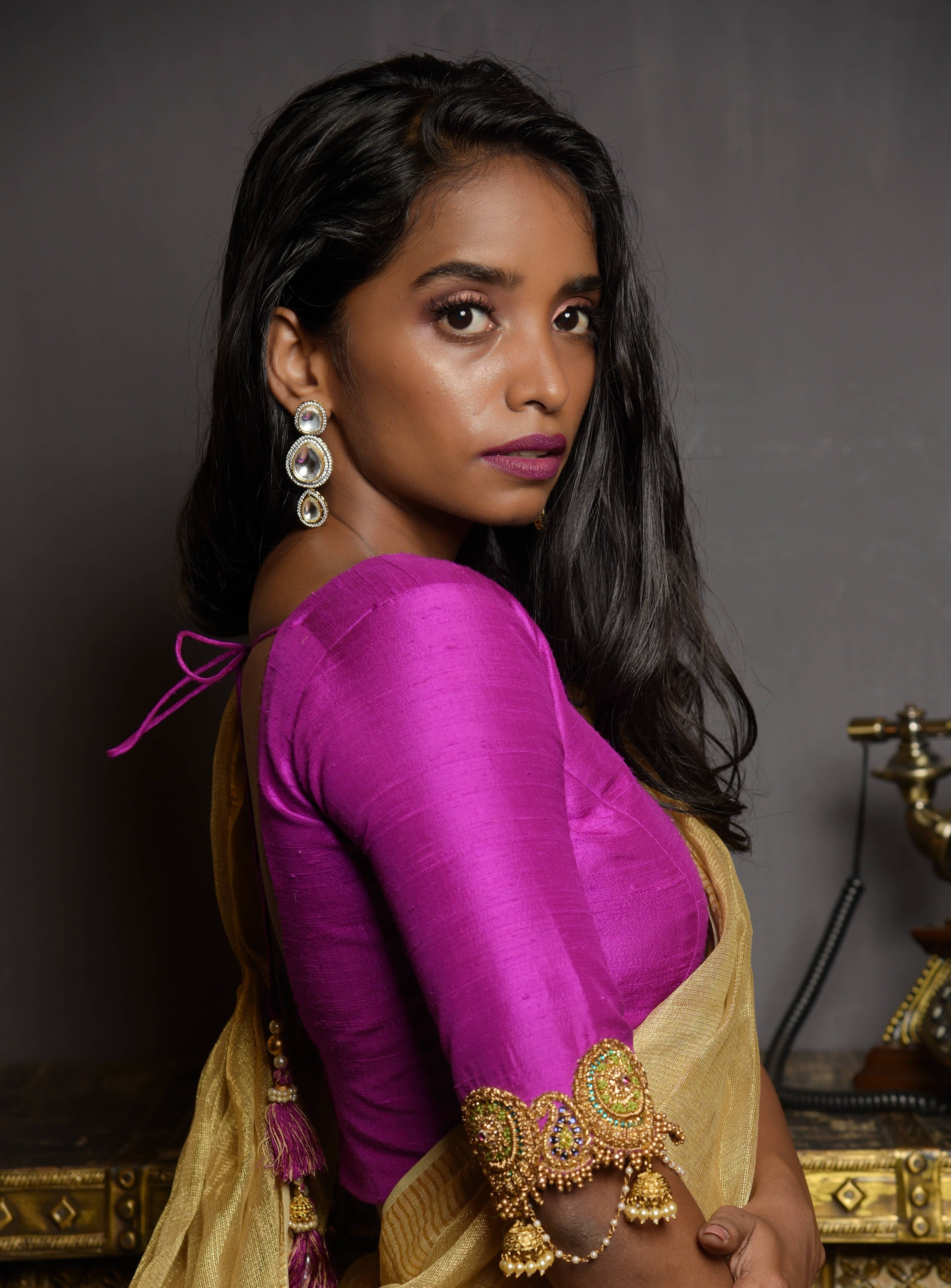 Model posing in a magenta raw silk blouse with zardozi and kemp embellishments on the edges of sleeves with cut work. The model has a dusky skin tone and has a tissue fabric draped like a dhoti so that the attention is on the blouse sleeve. She has turned her neck to the right and is looking straight at the lens.