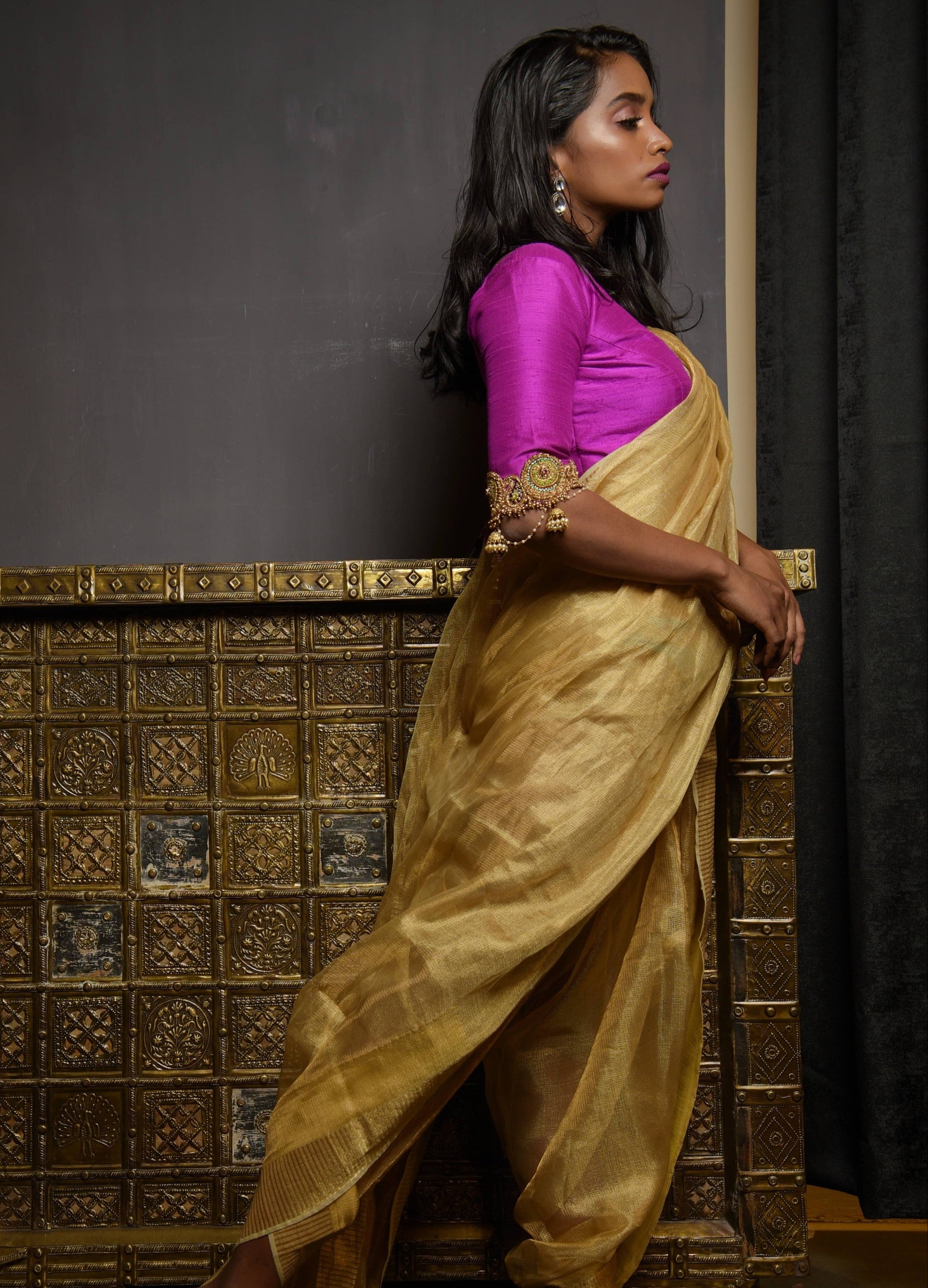 Model posing in a magenta raw silk blouse with zardozi and kemp embellishments on the edges of sleeves with cut work. The model has a dusky skin tone and has a tissue fabric draped like a dhoti so that the attention is on the blouse sleeve. Her side profile is fully captured.