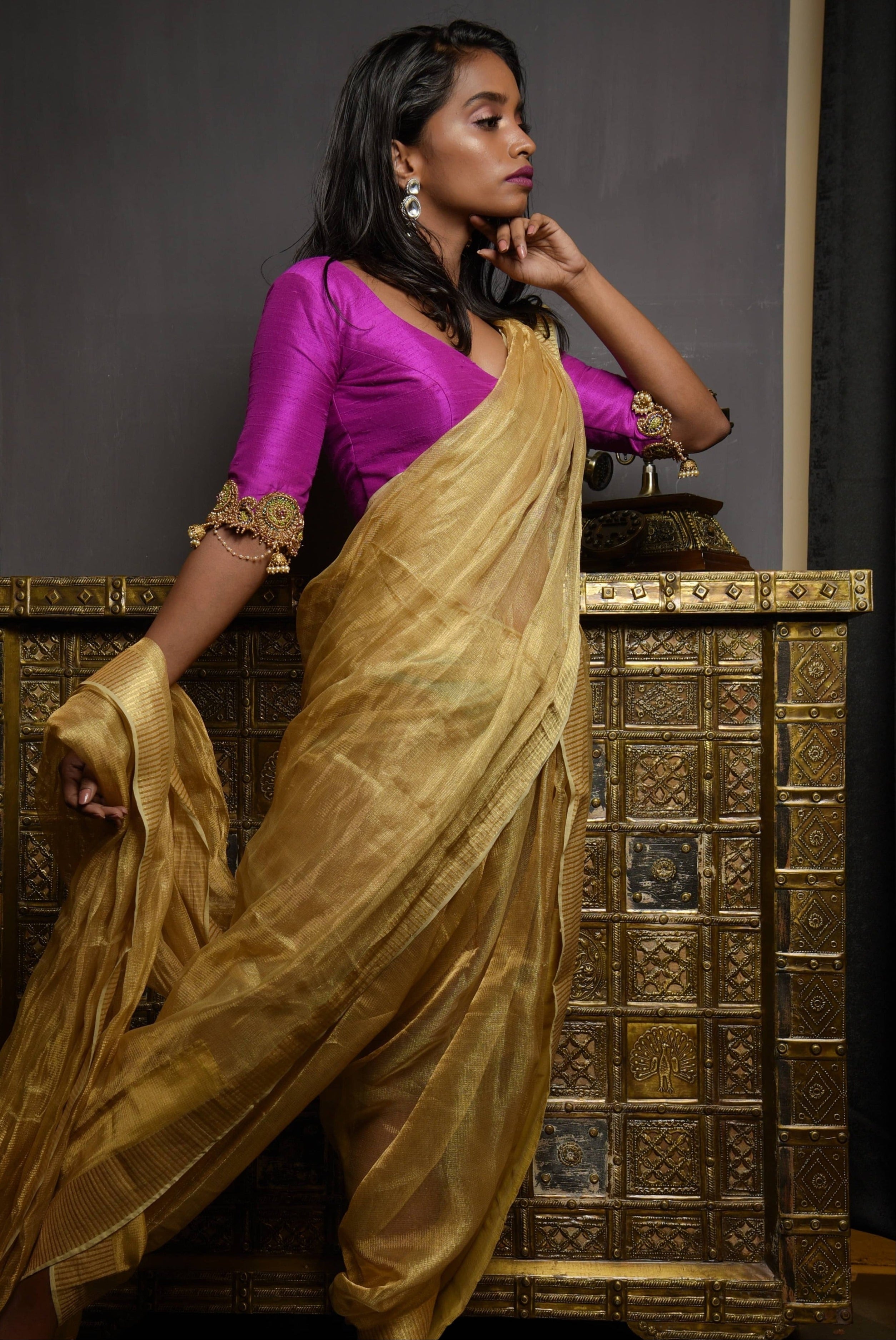 Model posing in a magenta raw silk blouse with zardozi and kemp embellishments on the edges of sleeves with cut work. The model has a dusky skin tone and has a tissue fabric draped like a dhoti so that the attention is on the blouse sleeve. She has kept her left hand undwer her chin and her side profile is captured fully.