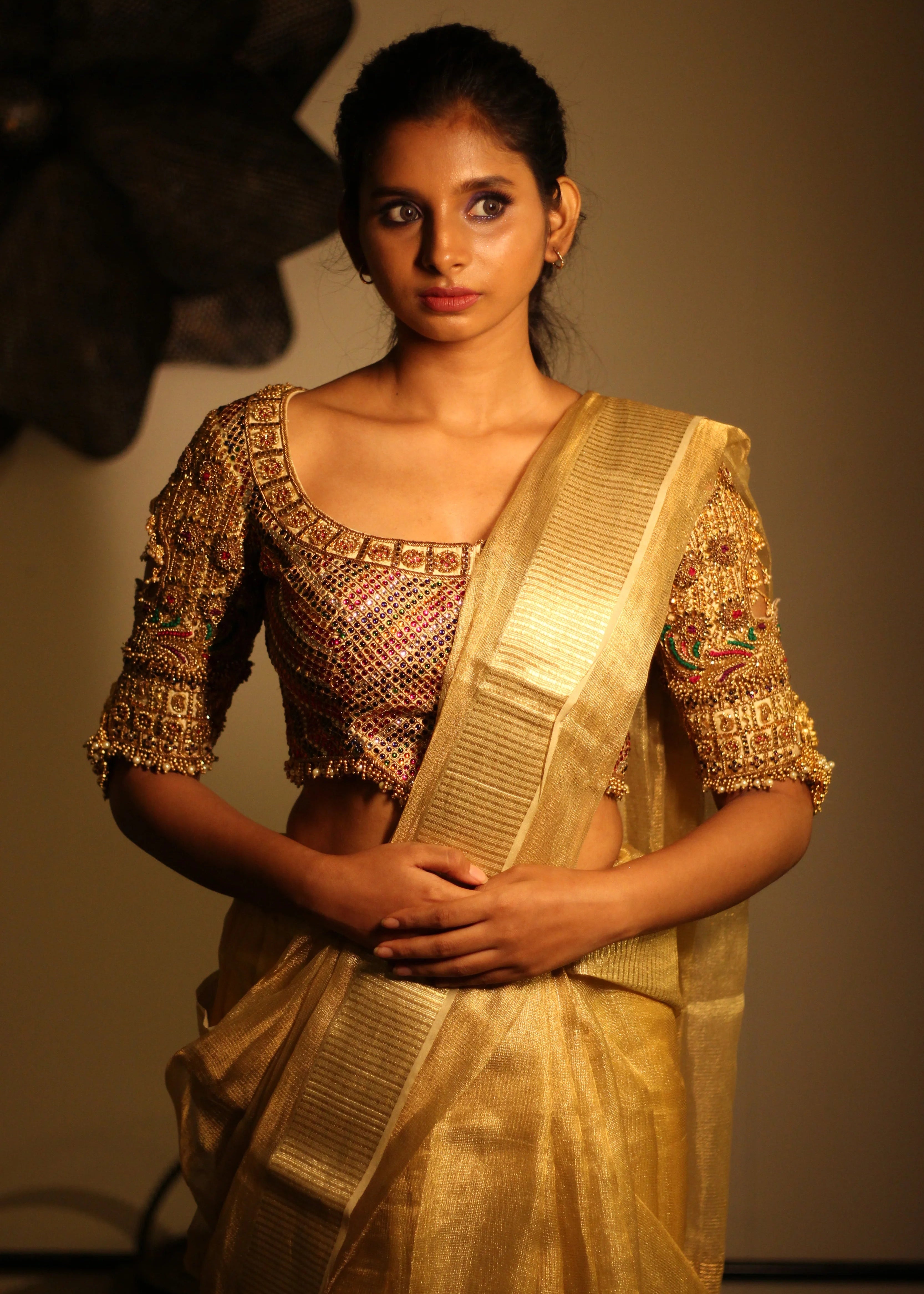 Model posing in a palanquin concept gold silk handcrafted blouse with heavy kemp and jhumki embeliishments and goddess pendants on sleeves.