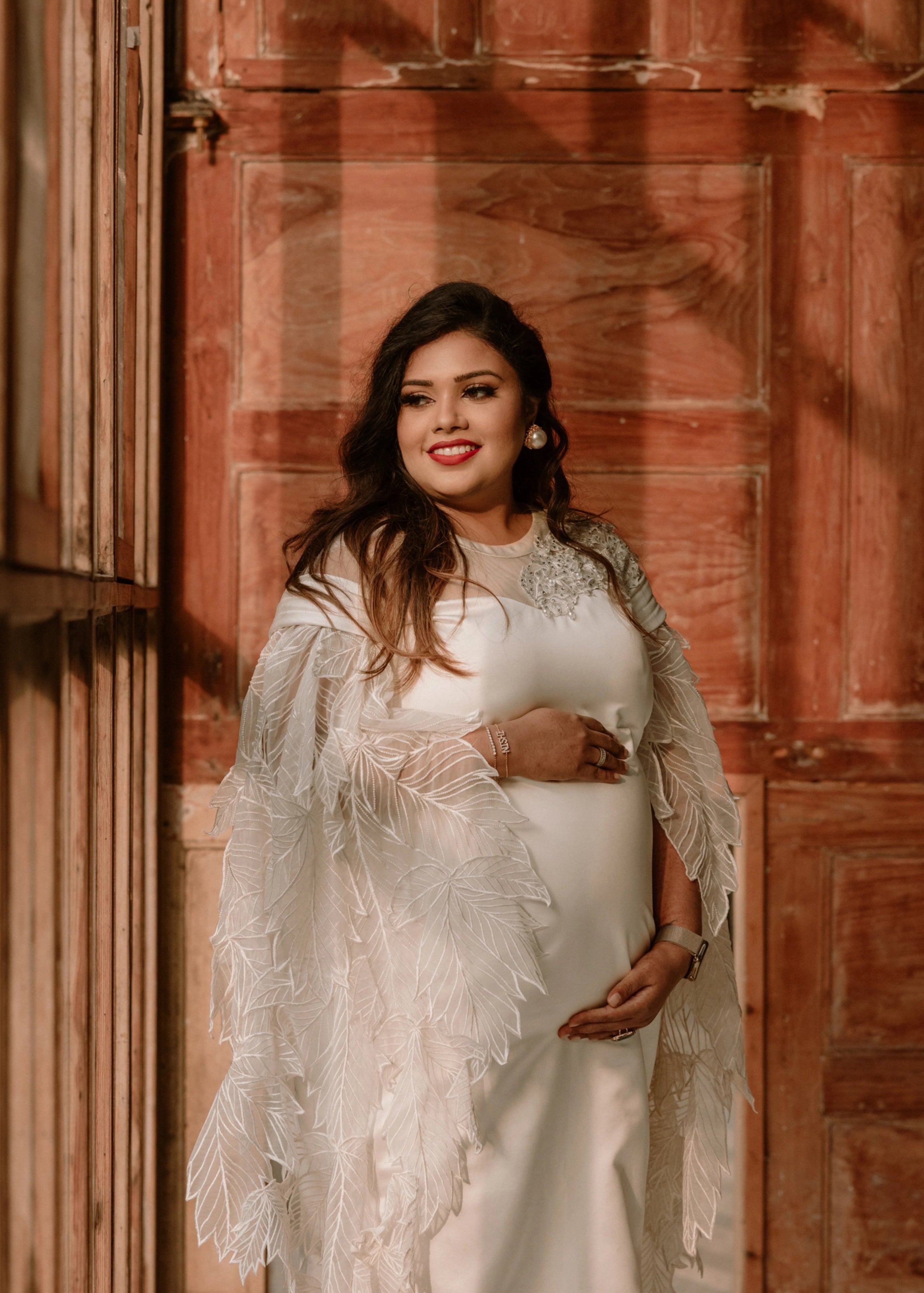 Mom to be Dr Nishitha posing in a white satin maternity gown, with leaf applique detailings by team Archana Karthick.