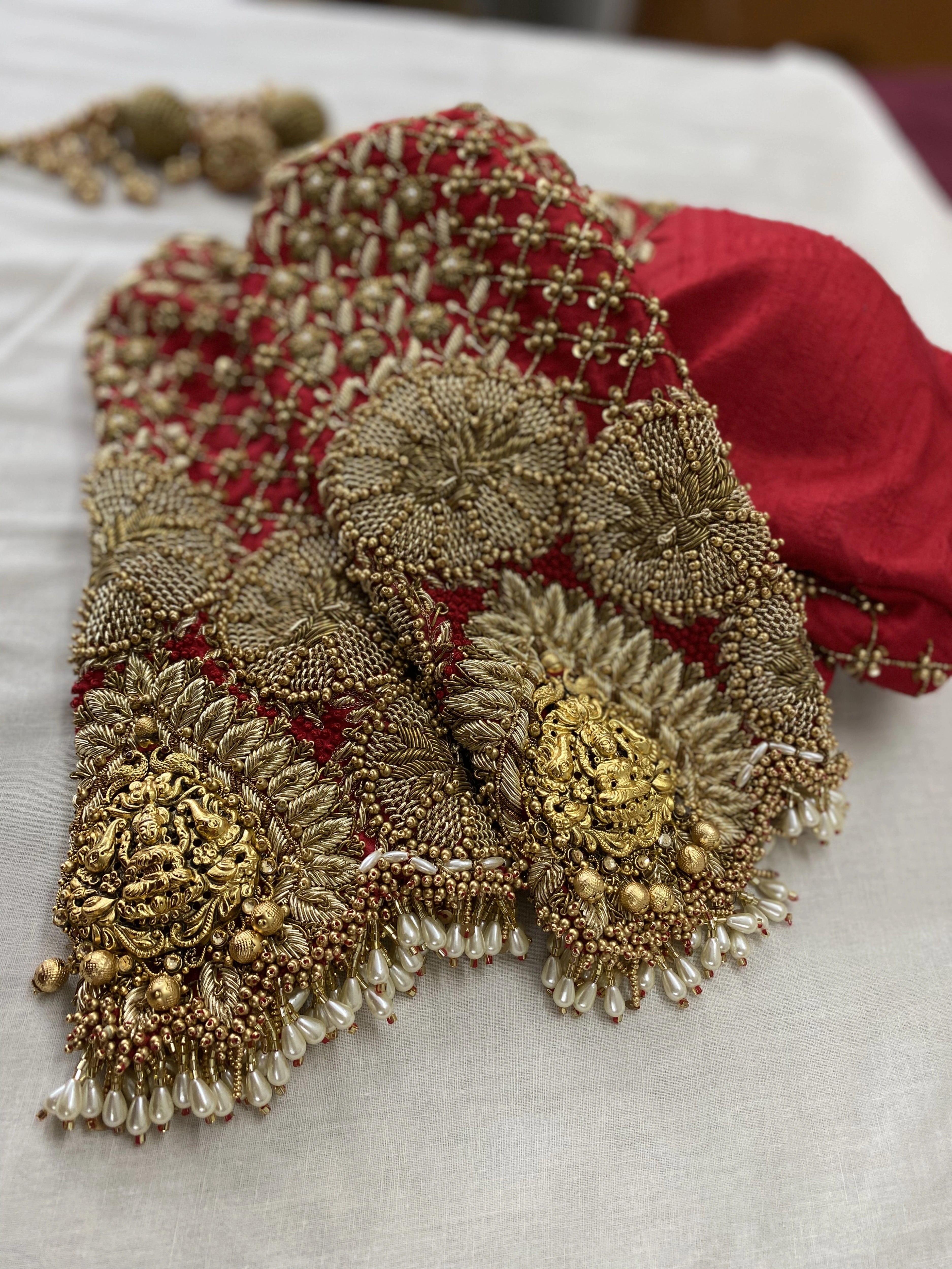Photo of a handcrafted red raw silk blouse embellished with zardozi, pearls, beads with goddess pendants on sleeves designed by Archana Karthick.