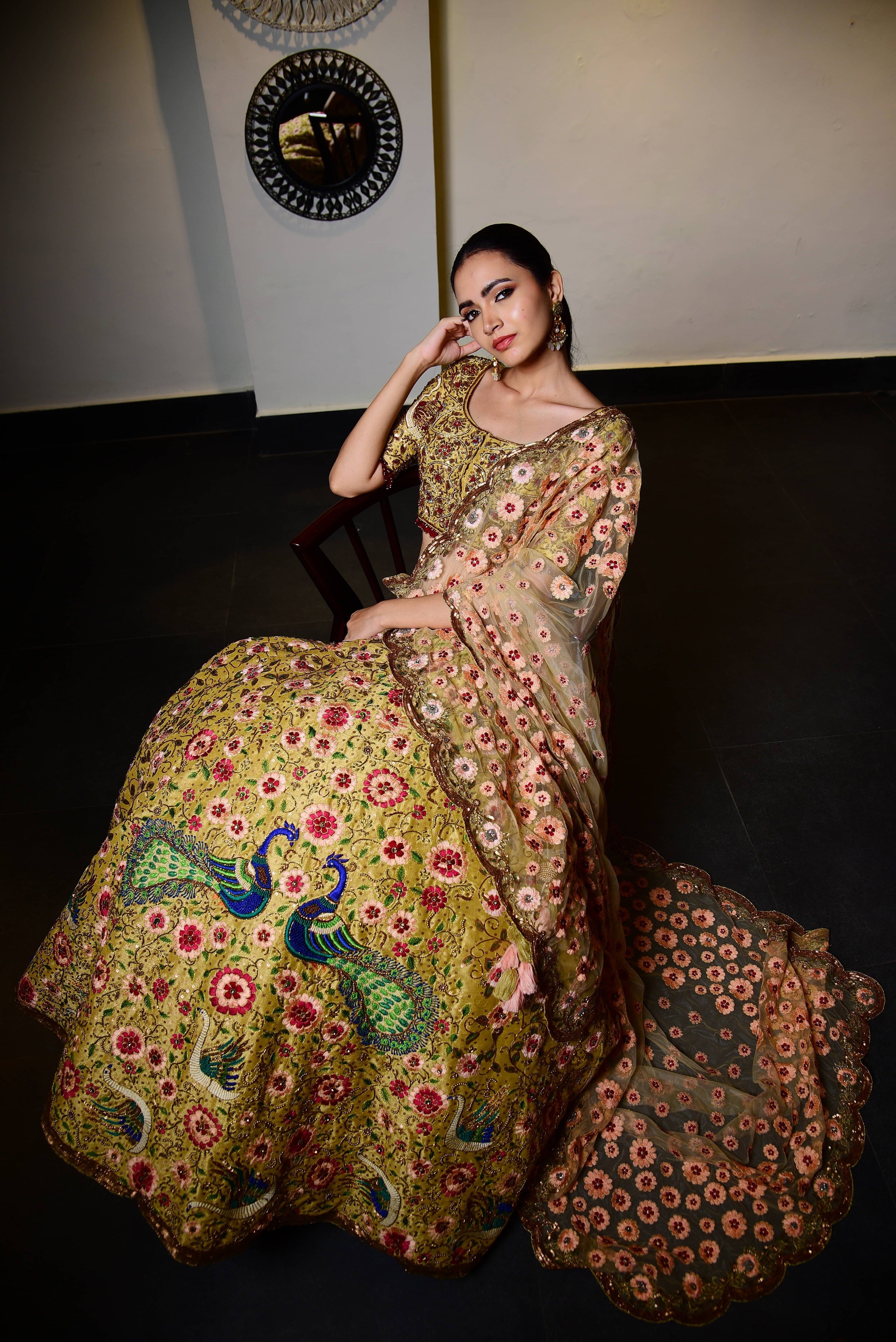 The model is posing in seated on a chair with her hand on her cheeks and a subtle smile where the side profile of the outfit revealed. This raw silk lehenga with heavy intricate resham, sequins, beads,stonework embellishments all over the lehenga, blouse and tulle dupatta.The beauty of the detailings on the dupatta or the veil is seen in detail.