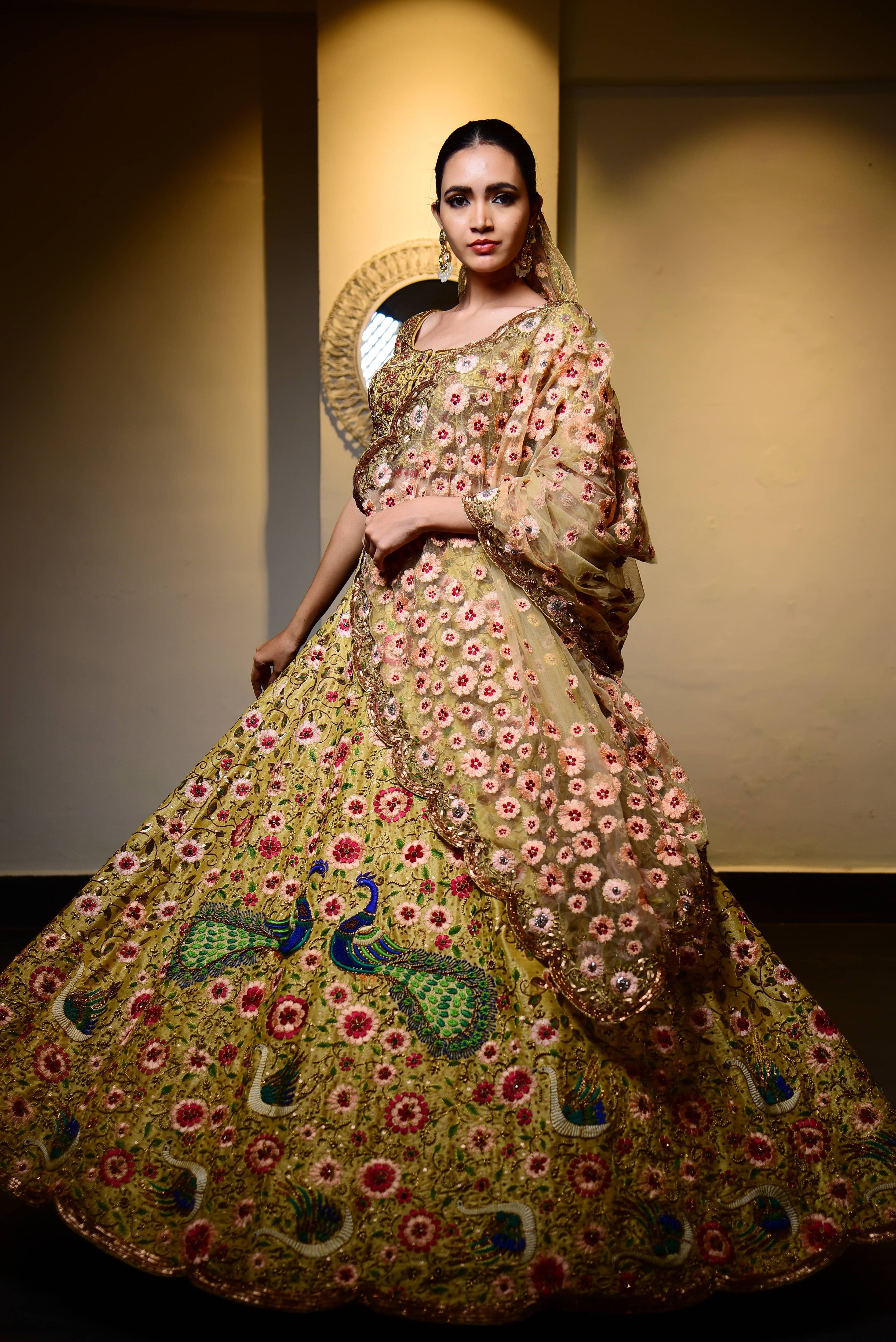 The model is posing where the complete front view of the outfit is seen and she is looking straight at the camera. This raw silk lehenga with heavy intricate resham, sequins, beads,stonework embellishments all over the lehenga, blouse and tulle dupatta.