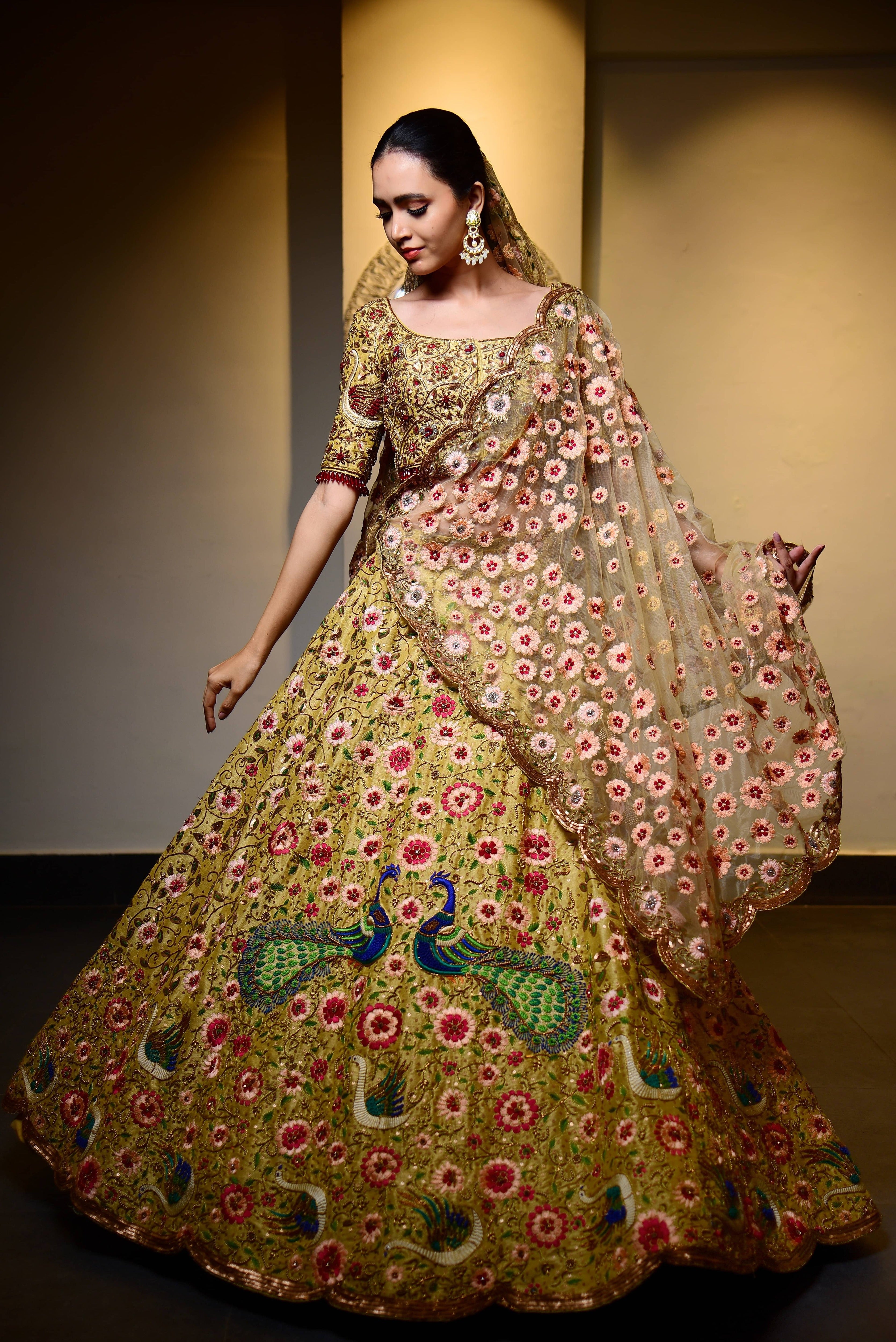 The model is posing where the complete front view of the outfit is seen and she is looking down making a shy gesture. This raw silk lehenga with heavy intricate resham, sequins, beads,stonework embellishments all over the lehenga, blouse and tulle dupatta w top half blouse front view