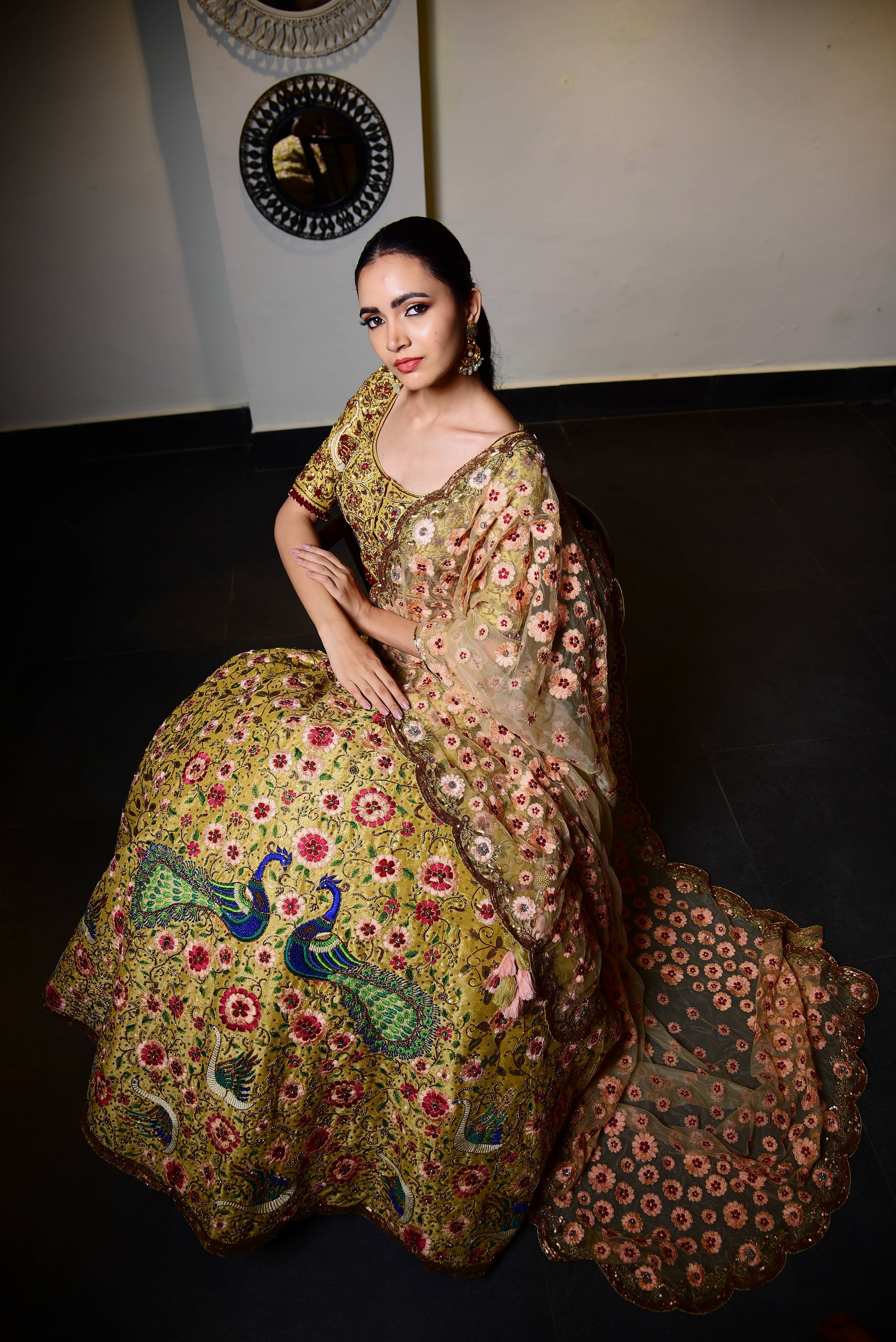 The model is posing in seated on a chair where the side profile of the outfit revealed. This raw silk lehenga with heavy intricate resham, sequins, beads,stonework embellishments all over the lehenga, blouse and tulle dupatta.The beauty of the detailings on the dupatta or the veil is seen in detail.