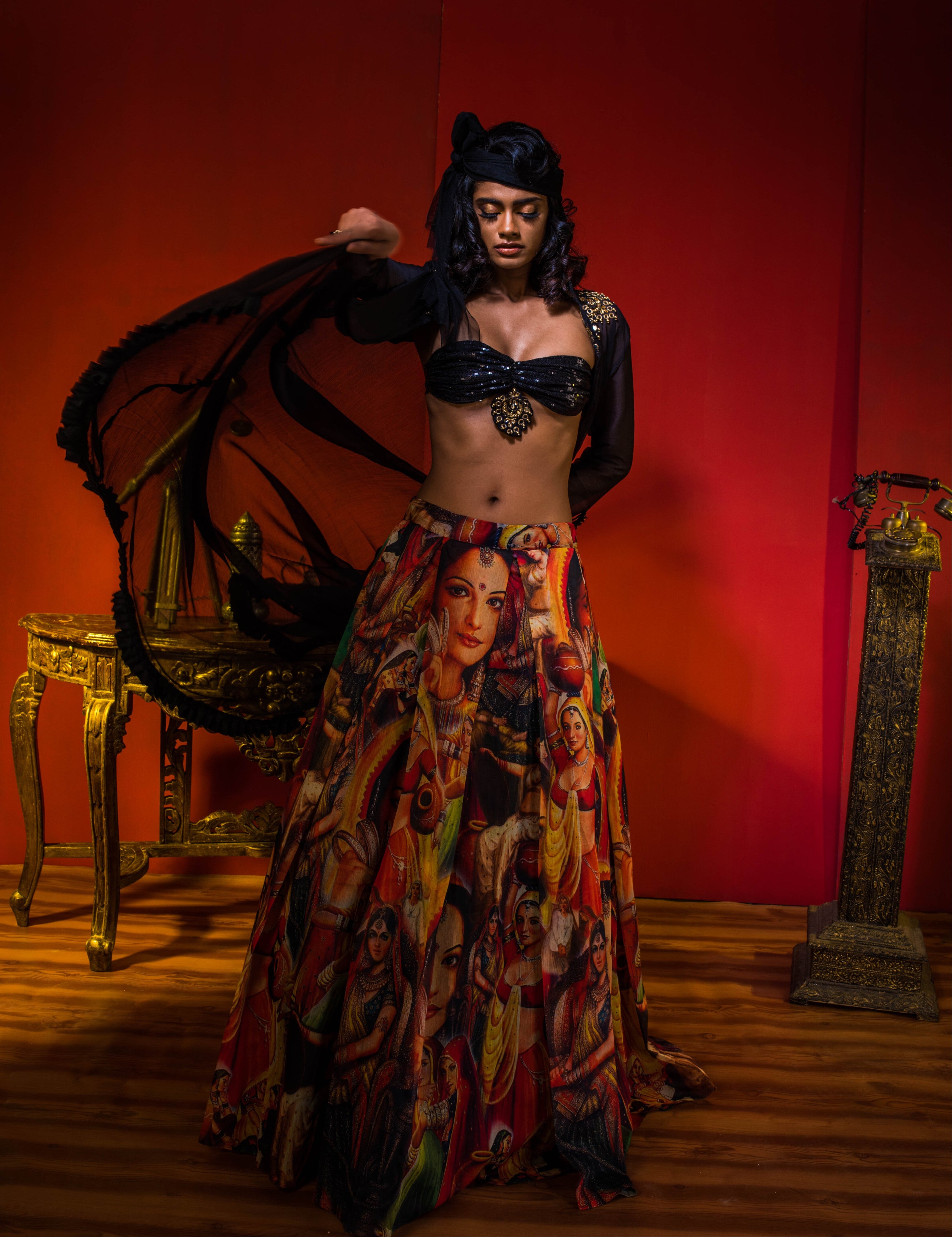 Supermodel Archana Akil Kumar in a heavily embellished black georgette bralette and long shrug paired with a printed colorful skirt looking down while waving the shrug to her right in an antique setting.