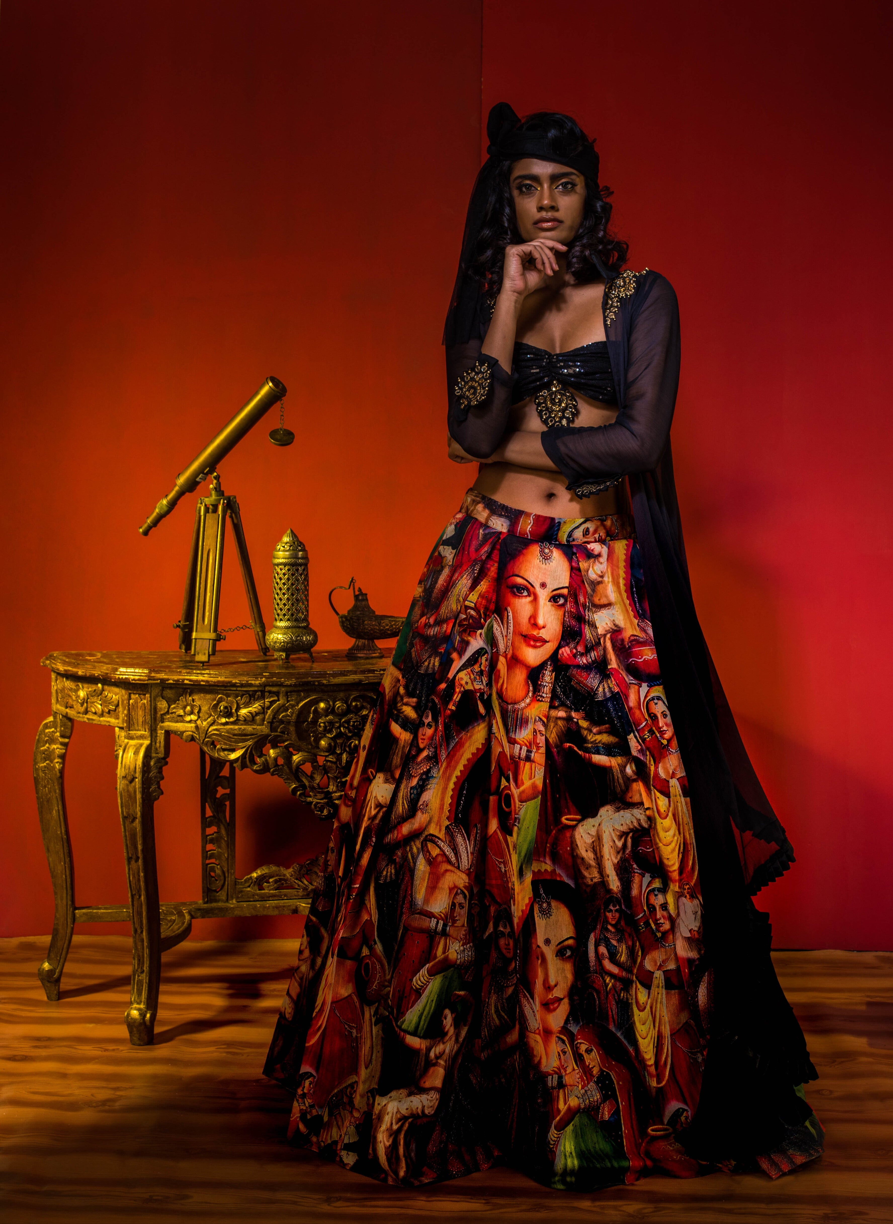 Supermodel Archana Akil Kumar in a heavily embellished black georgette bralette and long shrug paired with a printed colorful skirt looking staright into the lens with one arm bent and under her chin in an antique setting.