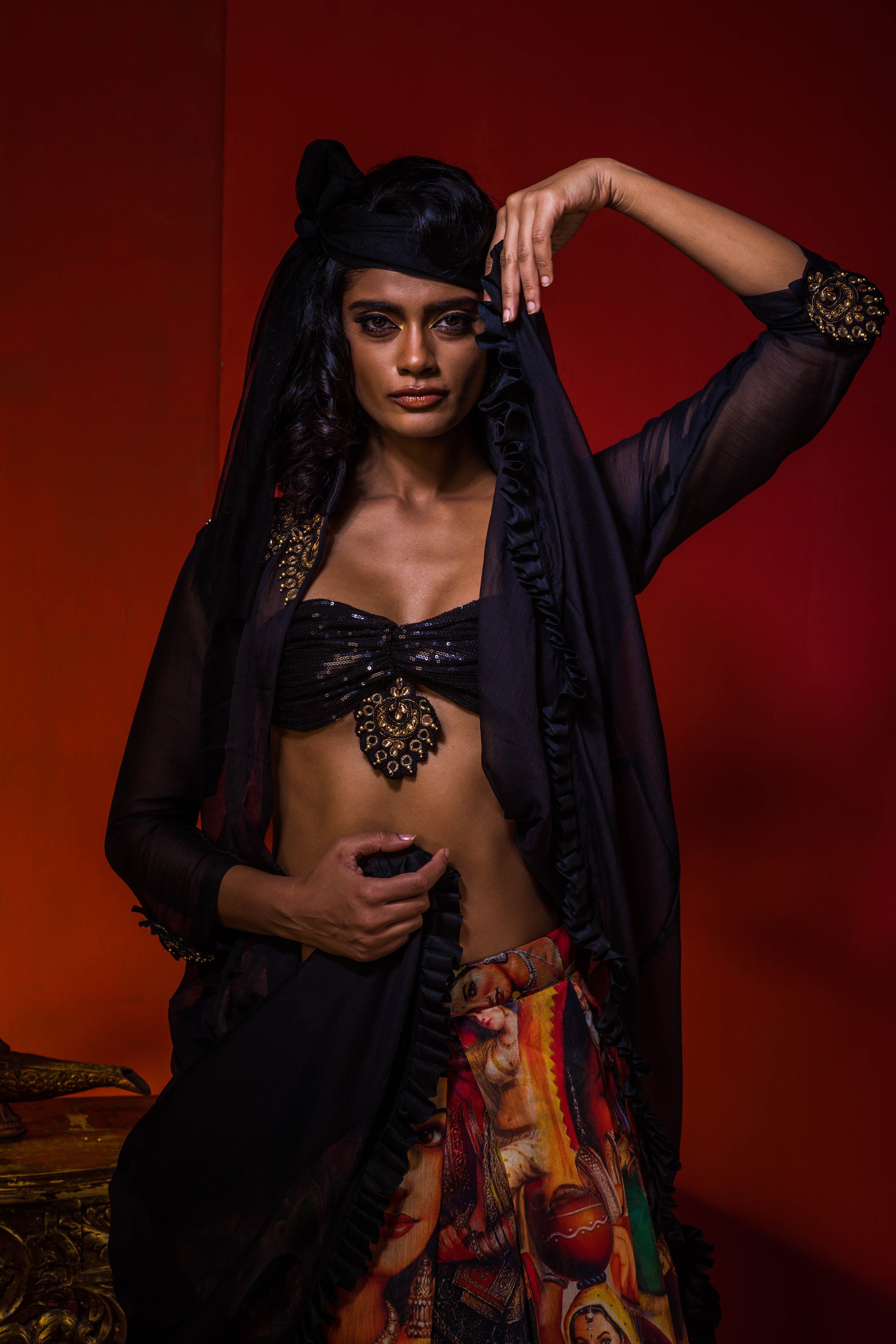 Supermodel Archana Akil Kumar in a heavily embellished black georgette bralette and long shrug paired with a printed colorful skirt looking staright into the lens with one arm lifting the shrug to her head and the other pulling the other end of the shrug to her waist in the front.