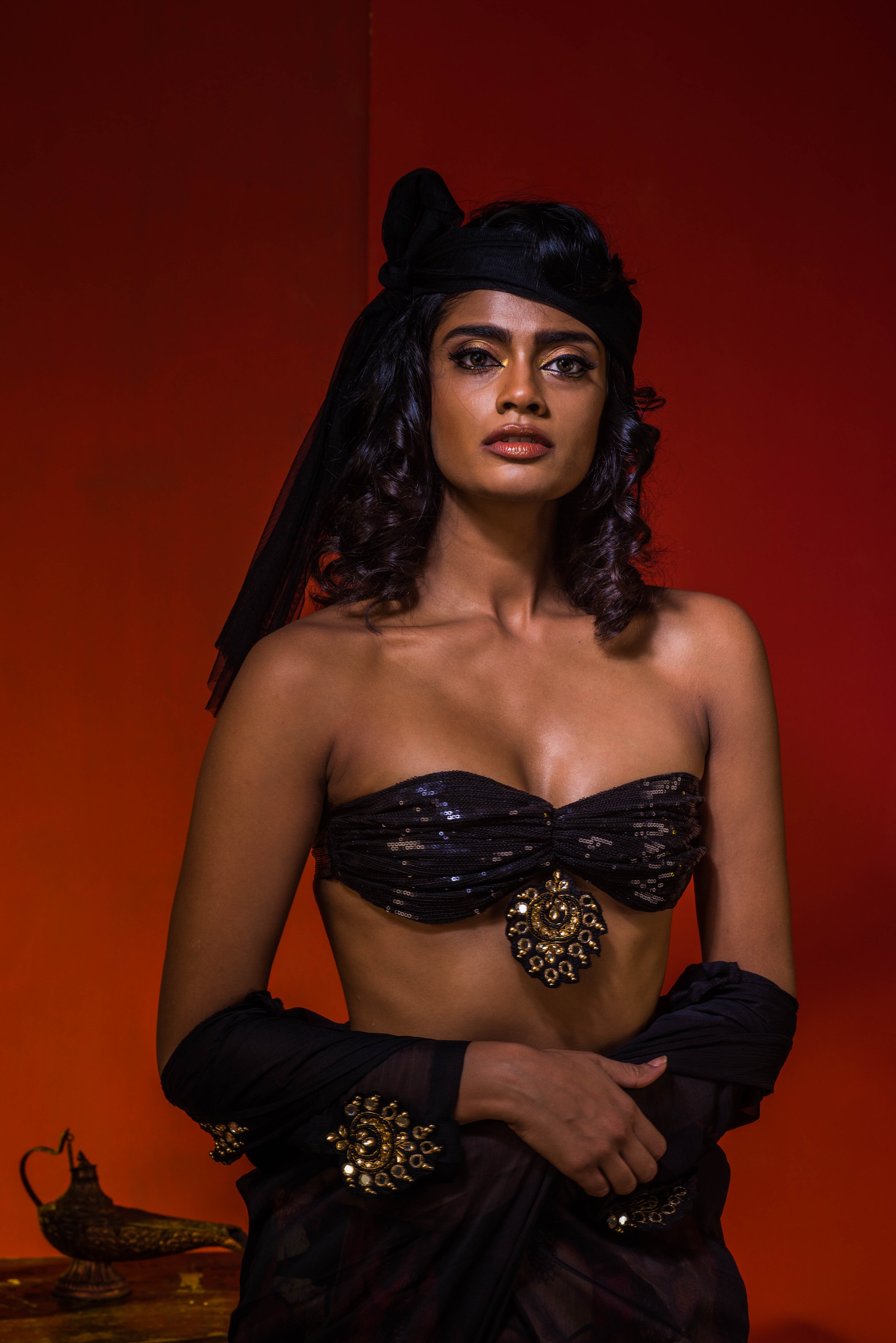 Supermodel Archana Akil Kumar in a heavily embellished black georgette bralette and long shrug paired with a printed colorful skirt looking staright into the lens with the shrug pulled down to her waist revealing the bralette and upper body.