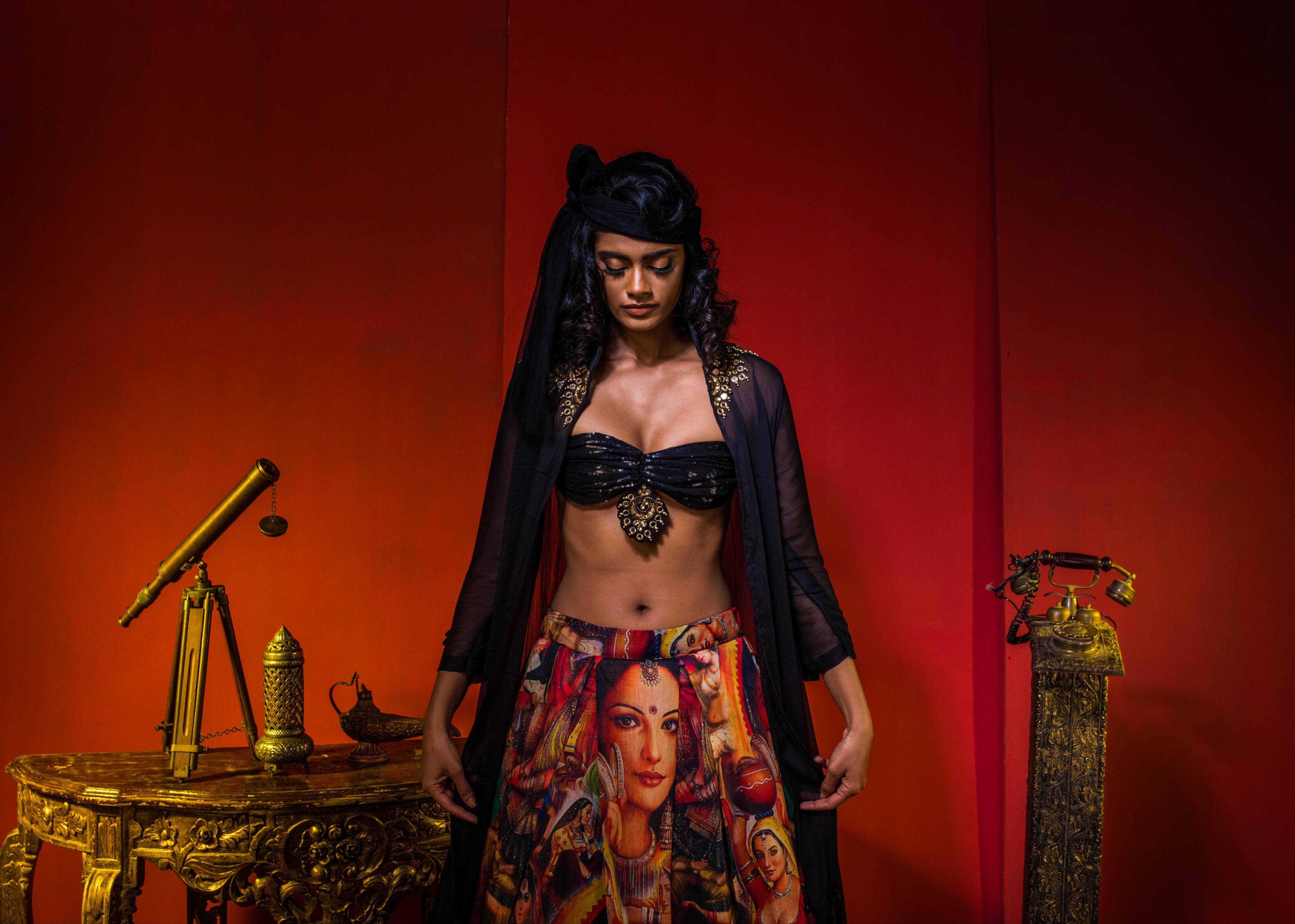Supermodel Archana Akil Kumar in a heavily embellished black georgette bralette and long shrug paired with a printed colorful skirt rlooking down with hands on sides in an antique setting.