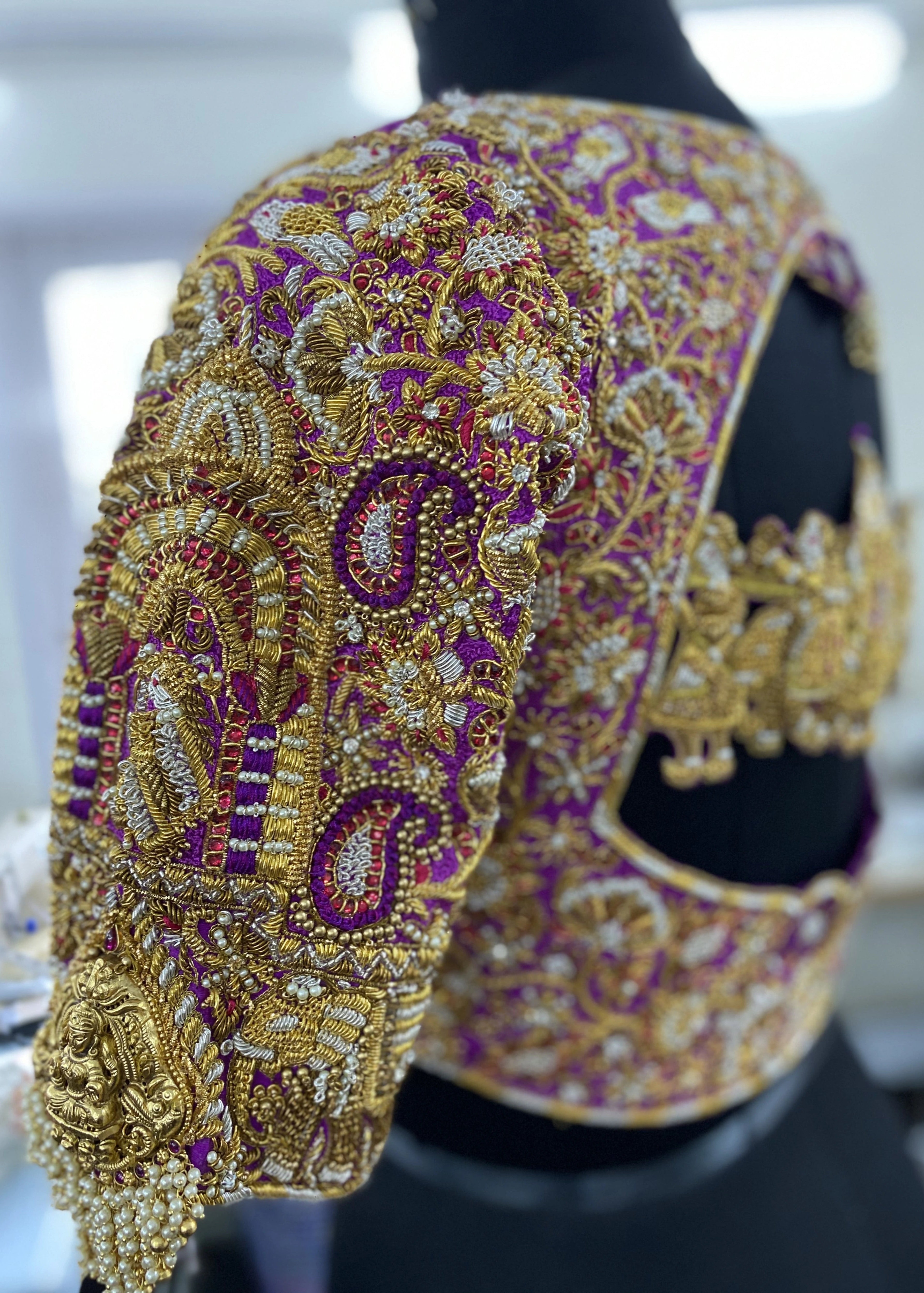 The sleeve detailing of a handcrafted palanquin concept magenta pink silk blouse with zardozi, kemp stones, pearls and resham knot work and goddess pendants on each sleeve. Designed by Archana Karthick