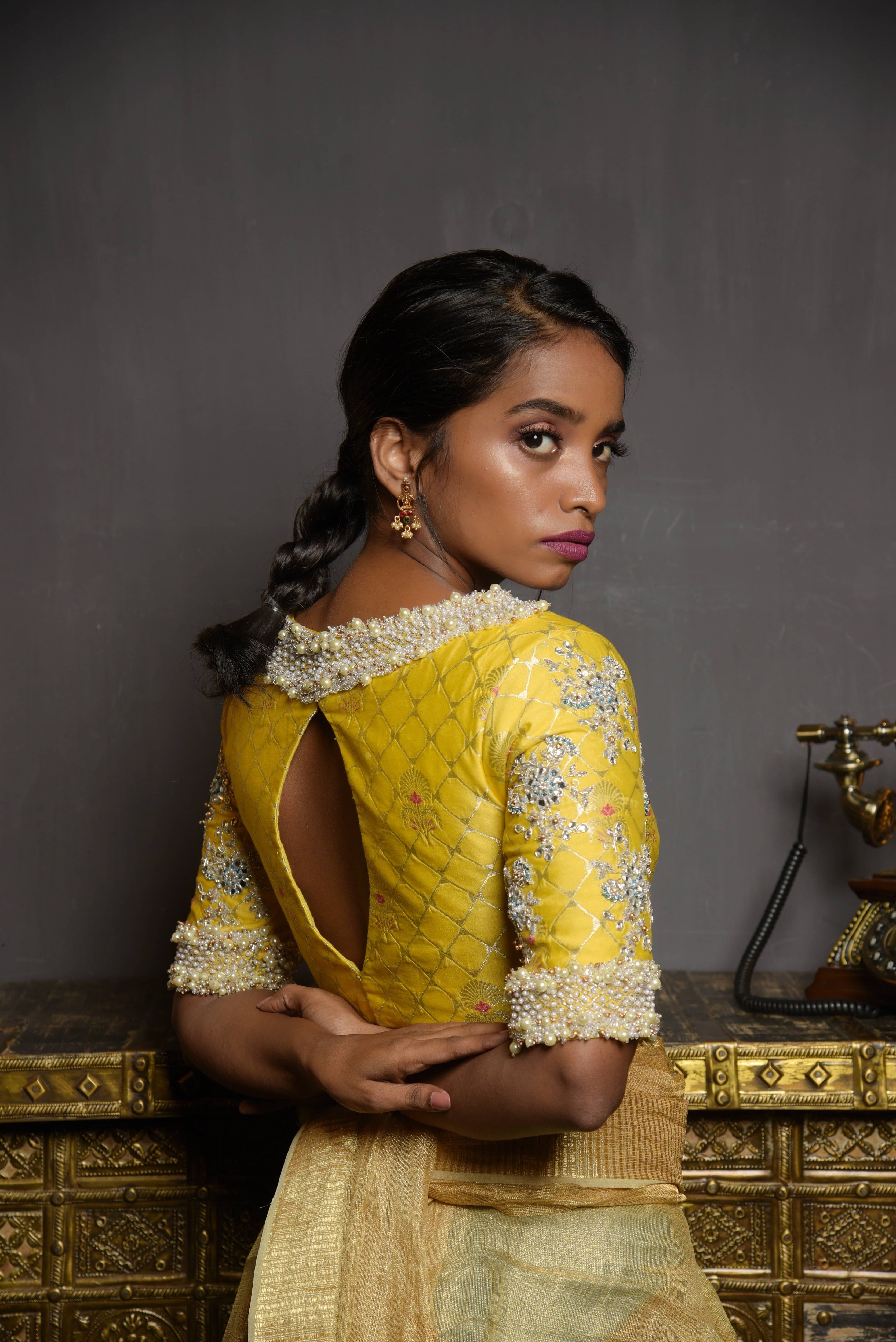 Model posing in a yellow banaras silk blouse with pearl embellishments on sleeves. The model looking behind her at the lens showing us the back neck blouse design.