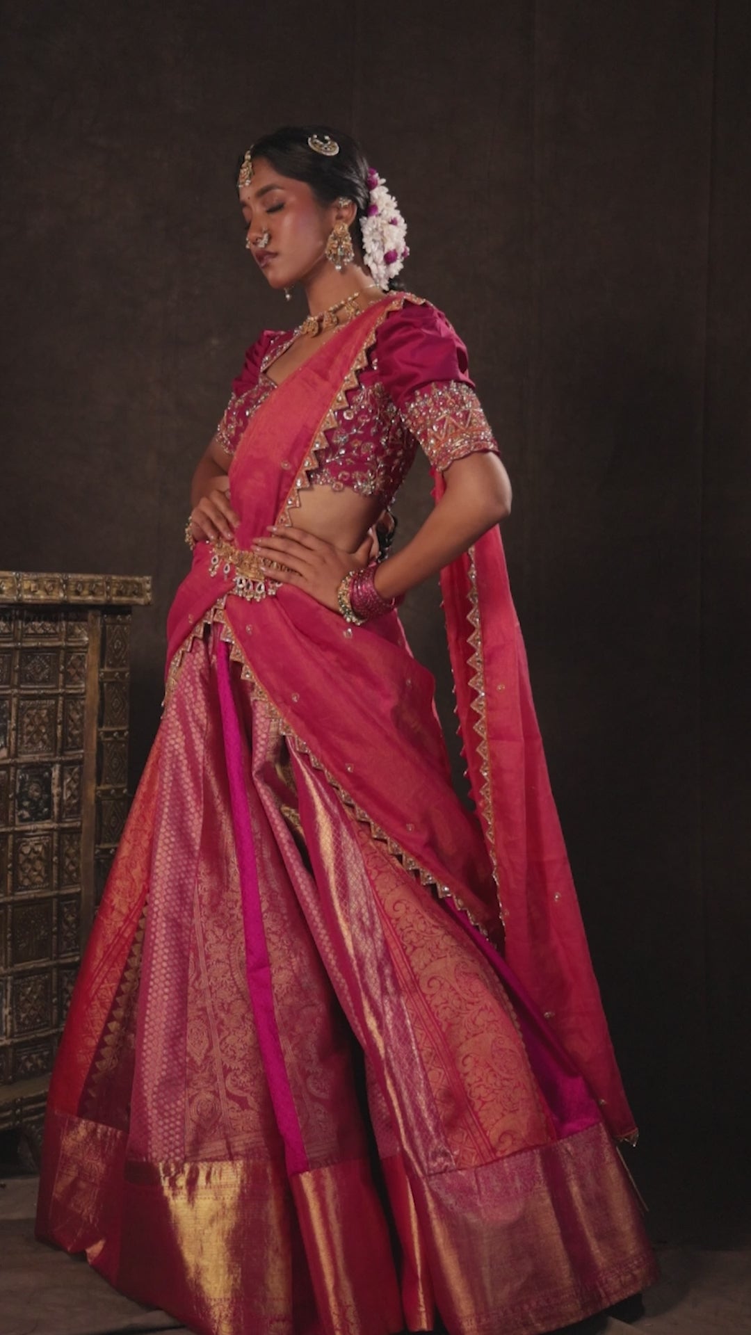 Model Gayathri Dhanasekar in a campaign video in a pink & orange silk half saree handcrafted with zardozi, cutdana (bugle beads), resham (silk thread), sequins and rhinestones. Team Archana Karthick completed the style with pink glass bangles, a ring, a choker, jhumkas, nose ring and a maang tikka.