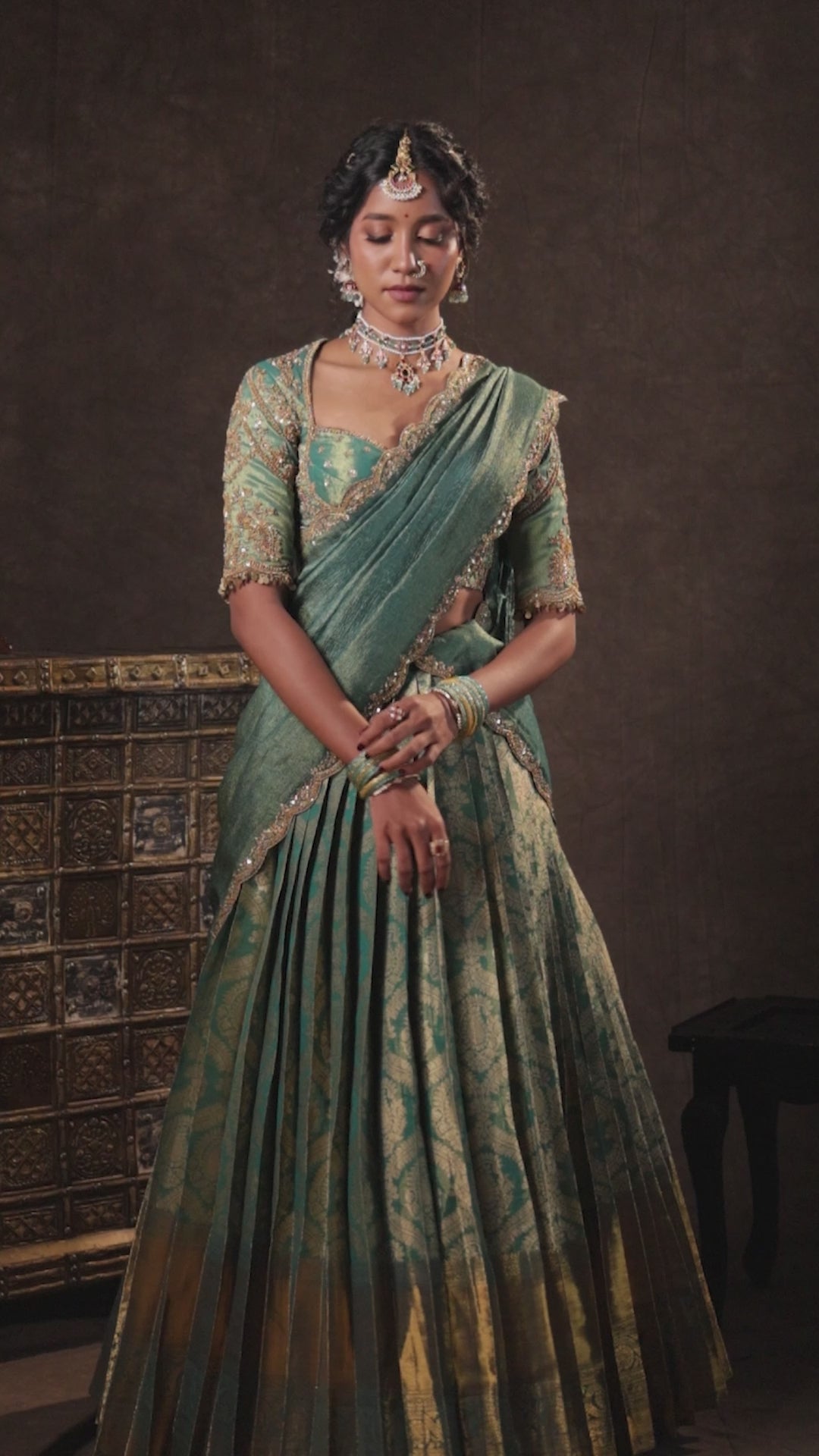 Model Gayathri Dhanasekar posing for a campaign video shoot in a teal half saree (silk blouse and skirt and tissue dupatta) handcrafted with zardozi, zari, cutdana, sequins and metal coin hangings. Team Archana Karthick completed the style with teal glass bangles, a ring, a choker, jhumkas, nose ring and a maang tikka.