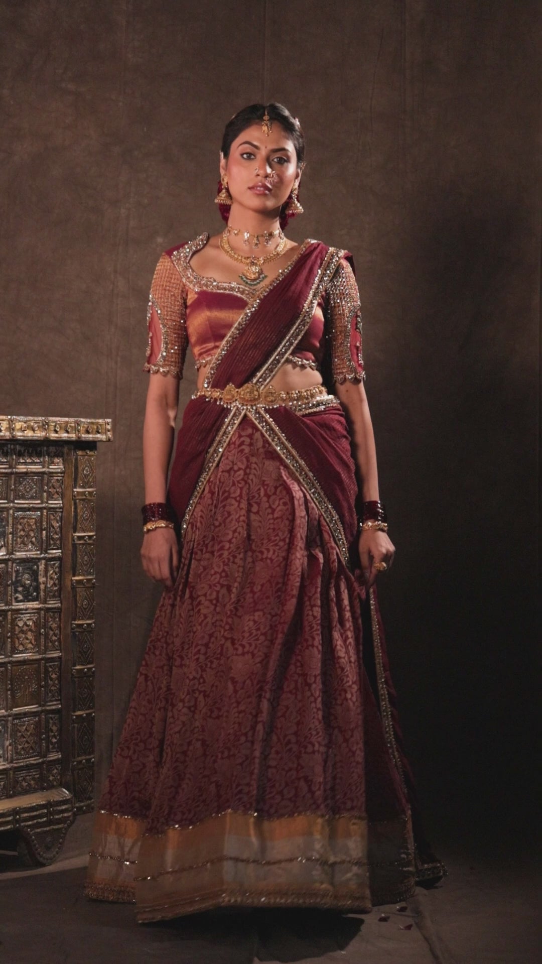 Model Dr Sree Advaita in a campaign video in a brownish maroon silk half saree set intricately handcrafted with zardozi, resham, cutdana, sequins & rhinestones. Team Archana Karthick completed the style with maroon glass bangles, a ring, jhumkas, nose ring and a maang tikka.