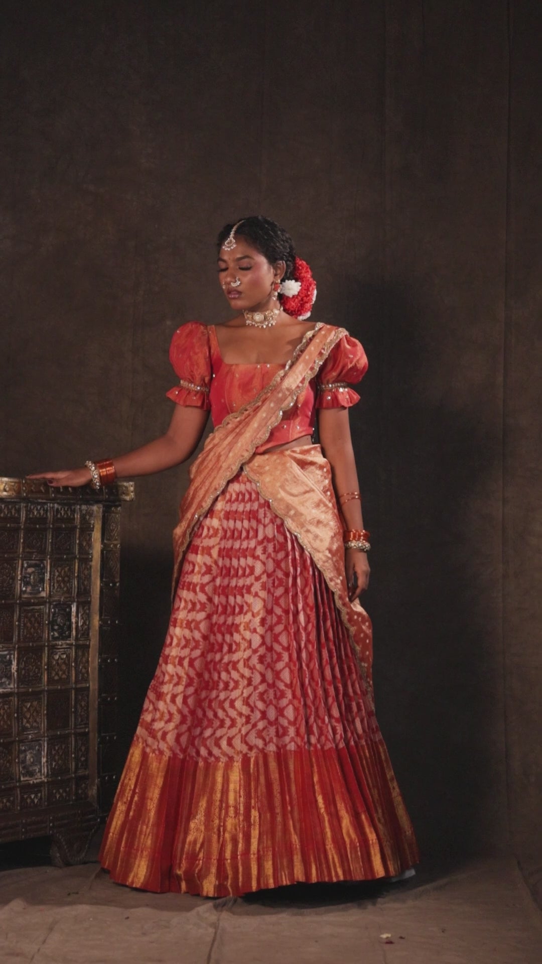 Model Ashika Crystal in a campaign video in a puff sleeved orange half saree set, handcrafted with zardozi, sequins, metal coin hangings and square cut stones. Team Archana Karthick completed the style with orange glass bangles, a ring, jhumkas, nose ring and a maang tikka.