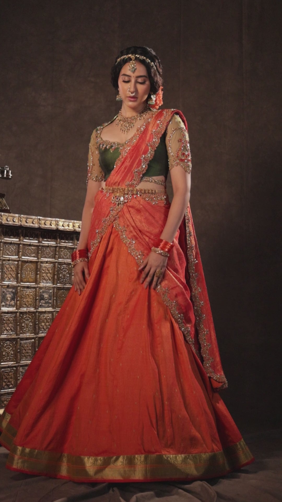 Actress Akshaya Hariharan for a campaign video in an orange & olive green silk half saree set with tulle sleeves. Handcrafted with zardozi, resham, cutdana, sequins & rhinestones. Team Archana Karthick completed the style with orange glass bangles, a ring, jhumkas, nose ring and a maang tikka.